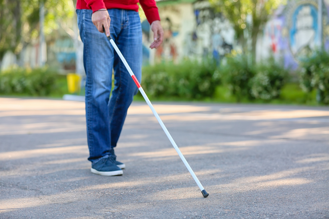 A person wearing jeans and tennis shoes holding a long white cane and walking on a sidewalk