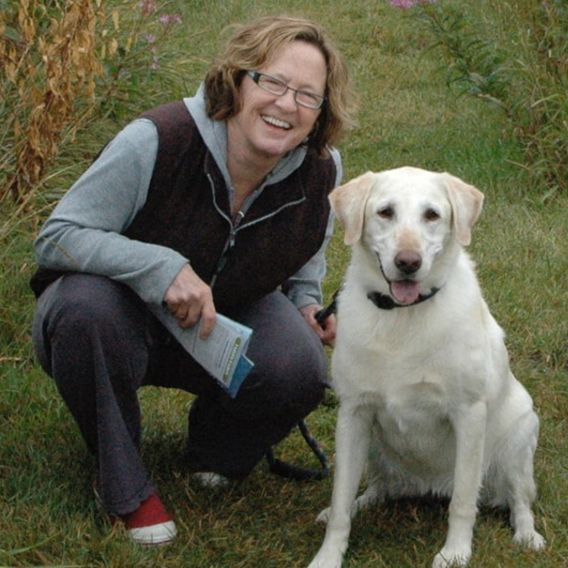 Photograph of Kari with her dog Clare. Kari is a white woman with shoulder length reddish brown hair. She is smiling and kneeling and holding on to Clare's leash. Clare is creamy colored therapy dog and one of the main characters in Kari's book Adalyn’s Clare.